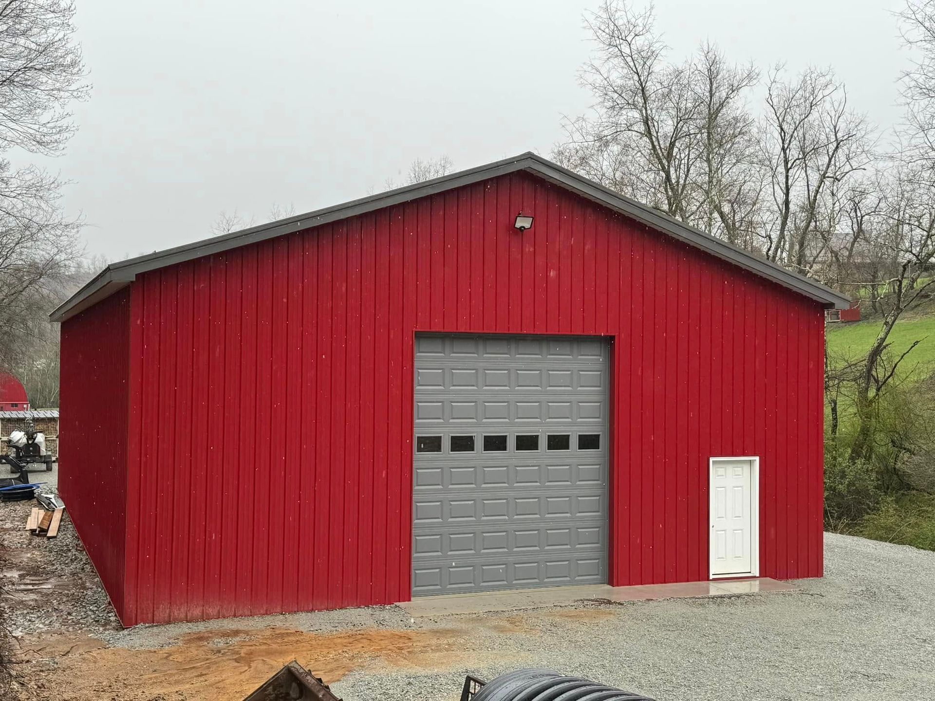 Red metal barn with a gray garage door and a white door to the side, set on gravel.