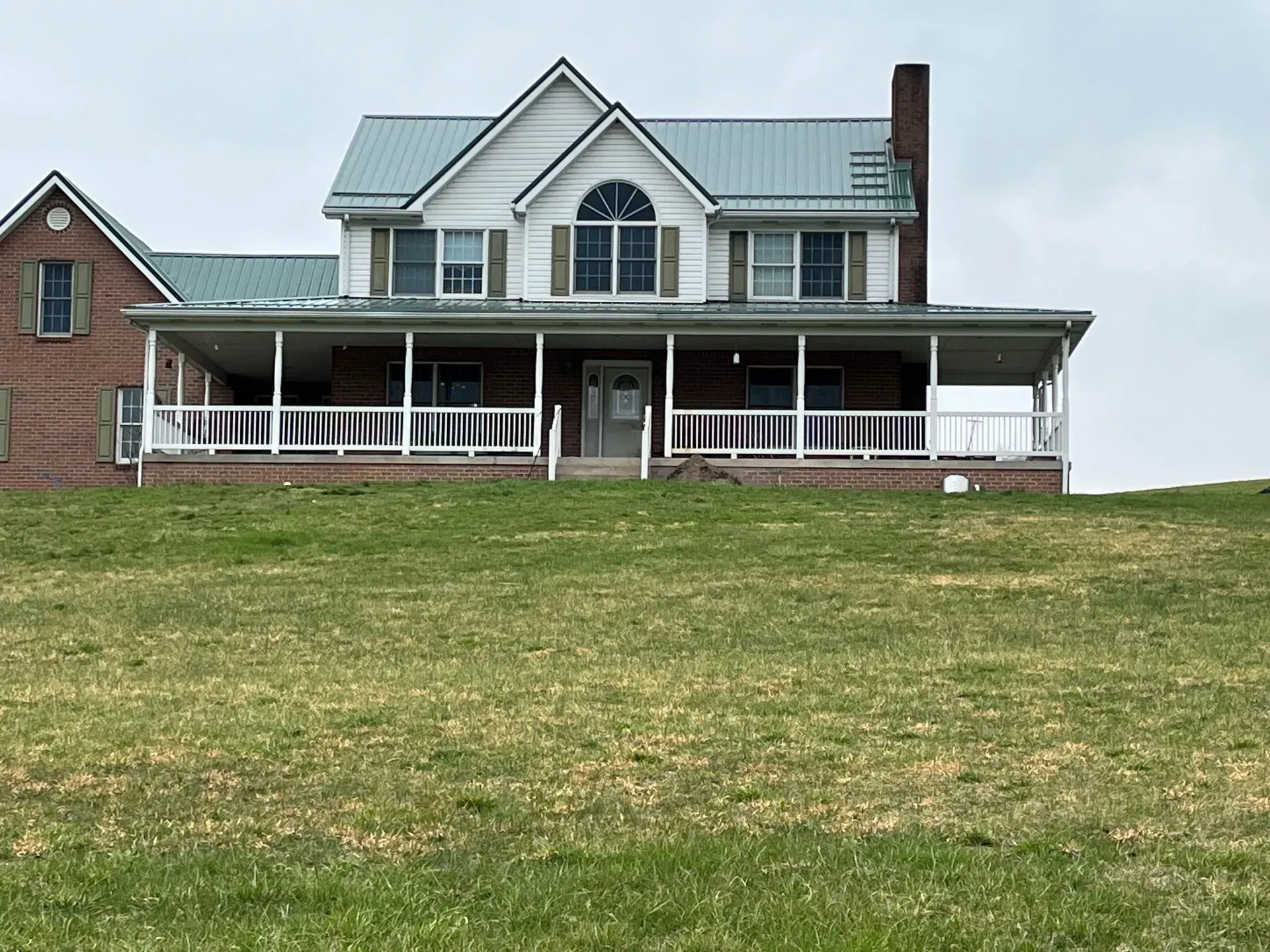 Farmhouse with a green metal roof and a wraparound porch on a grassy hill under an overcast sky.