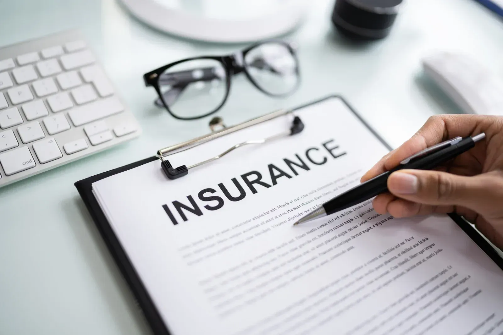 Person holding pen over an insurance document on a clipboard at a desk, with glasses, keyboard, and mouse visible.