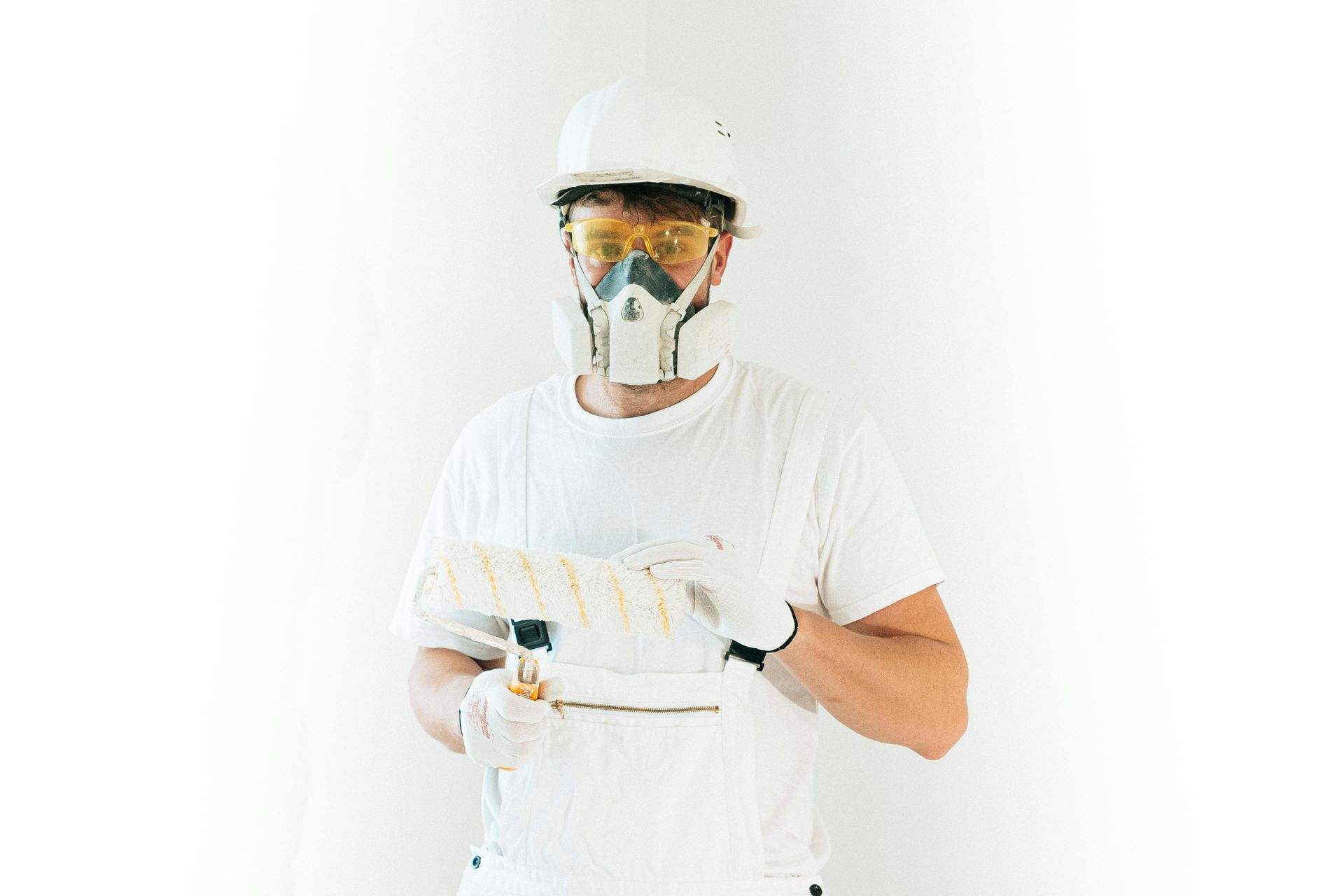 Person in white protective gear, helmet, mask, and goggles holds a paint roller against a white background.
