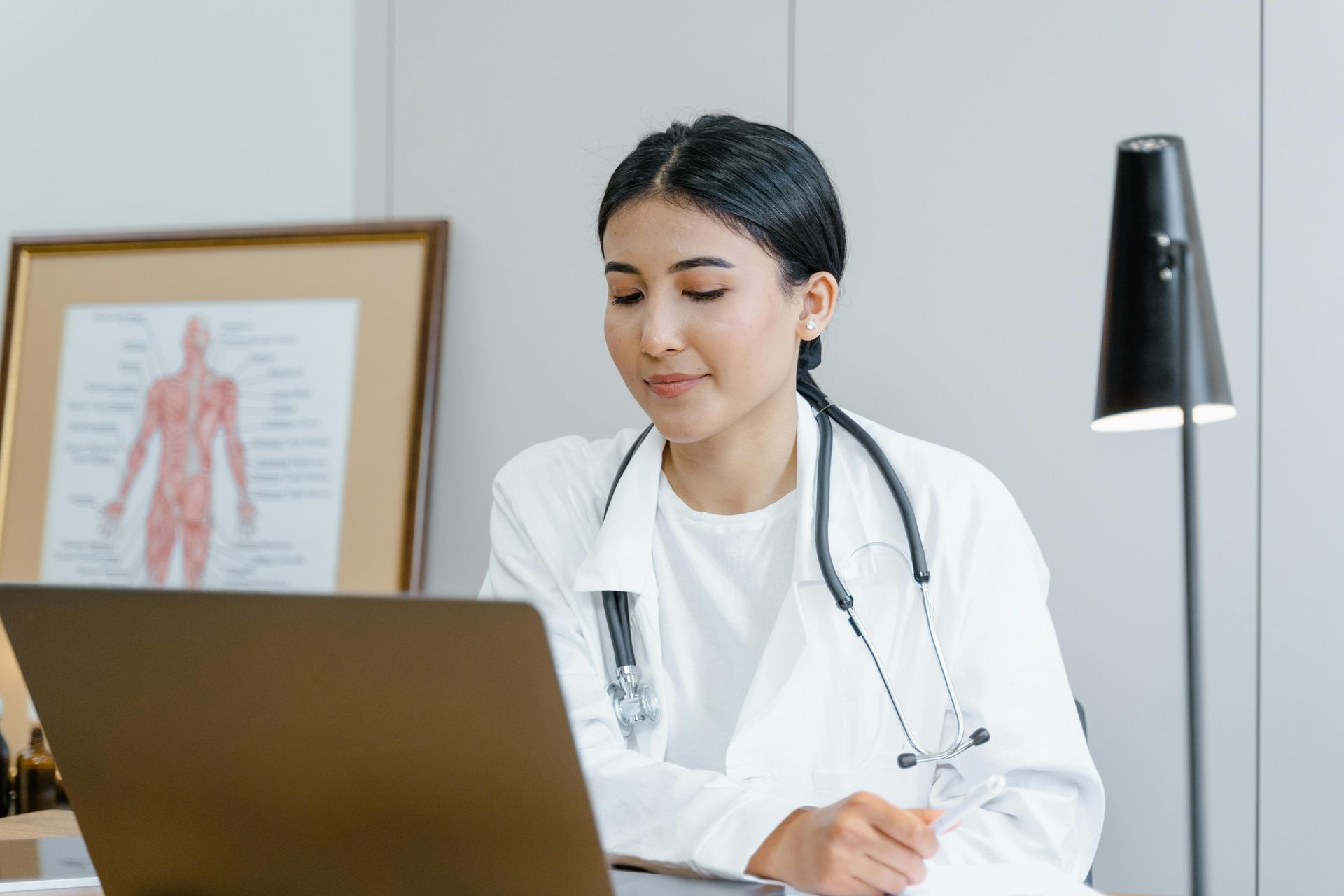 A medical professional in a white coat with a stethoscope around their neck looks at a laptop in an office.