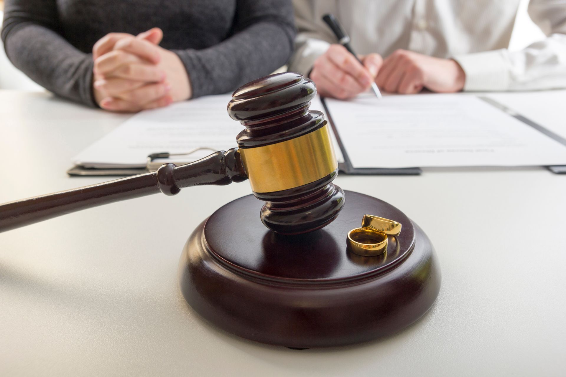 Gavel and wedding rings on desk with two people at table signing documents.