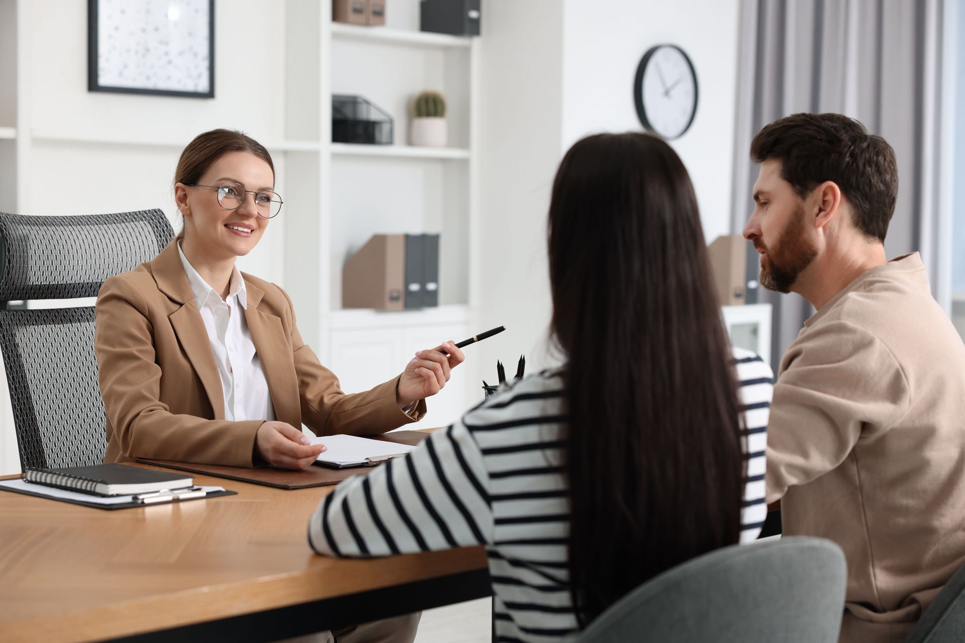 Woman in a blazer consults with a couple at a table, pointing at paperwork; office setting.