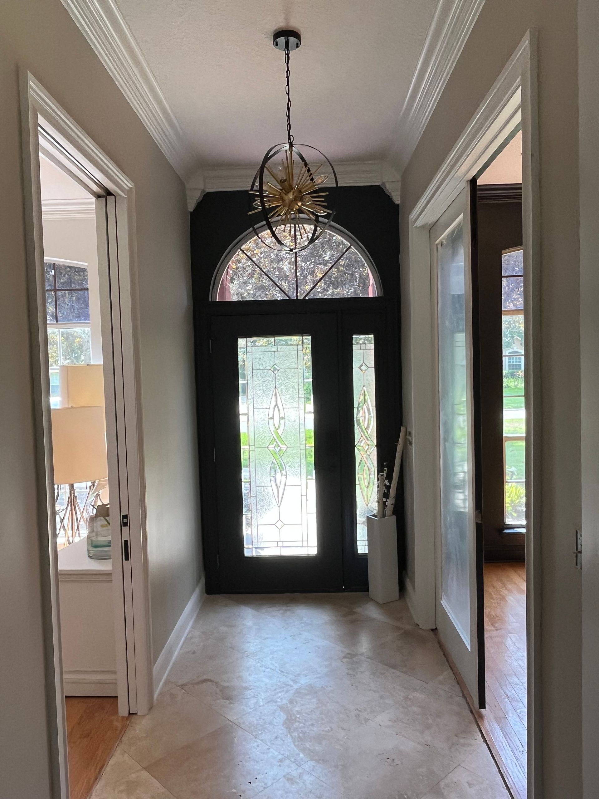 Narrow hallway with dark front door, chandelier, white walls, and side rooms opening off the corridor