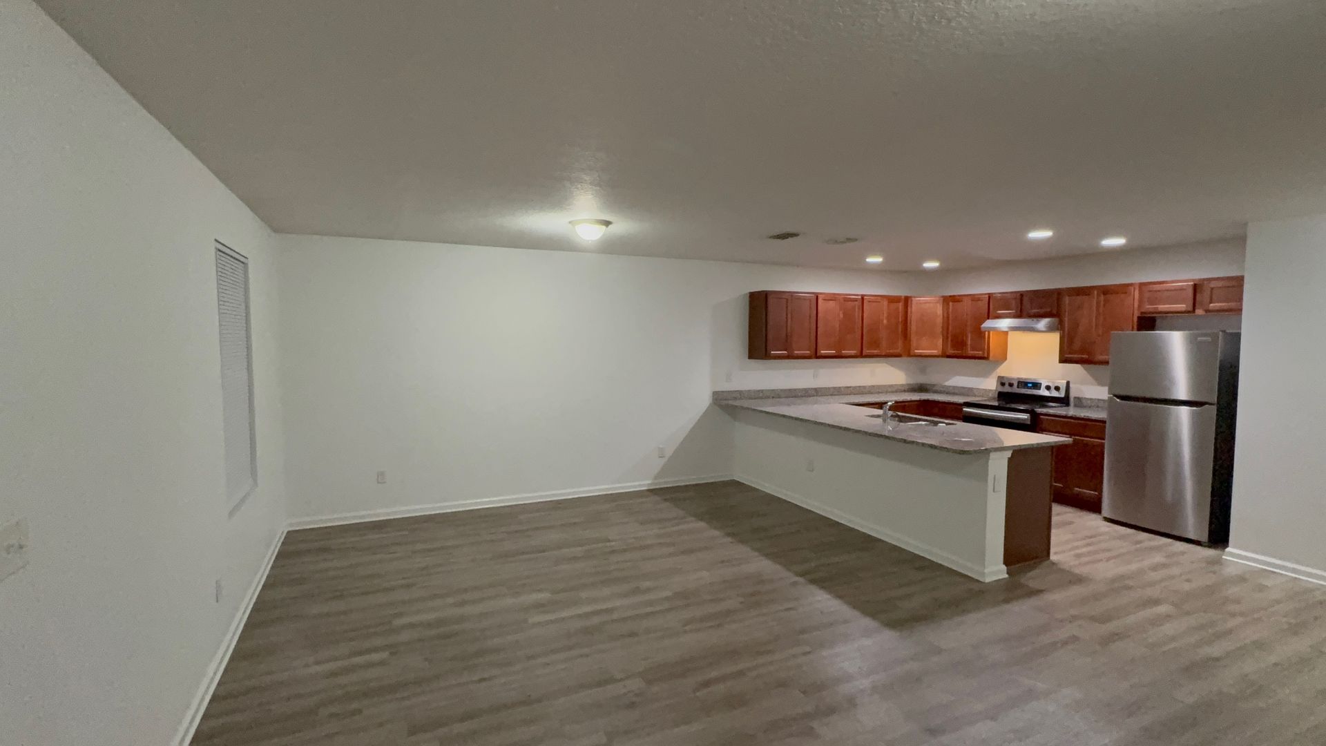 Empty open-plan living room with modern kitchen, white walls, wood floors, and stainless steel fridge.