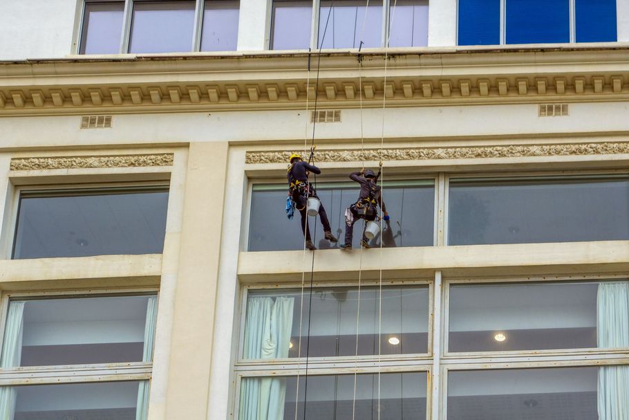 Two window washers suspended on ropes outside a beige building facade