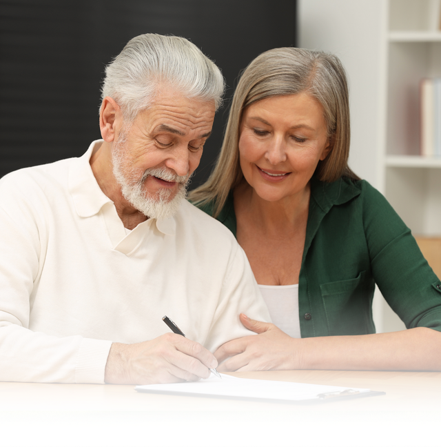 Two people sit together, one signing a document while the other watches closely in a bright indoor setting.