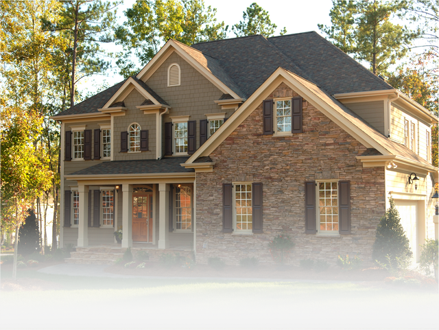 A large, two-story house with a stone facade, dark shutters, and a shingled roof, surrounded by trees.