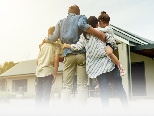 A family stands with their arms around each other, looking toward a house, seen from behind.