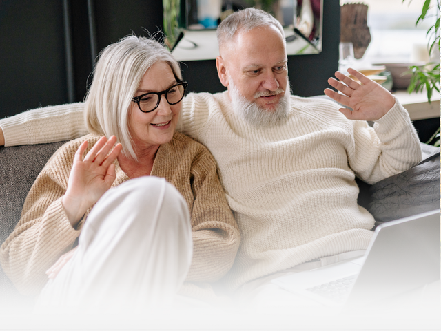 Two people in sweaters sit on a couch, smiling and waving while video calling on a laptop.