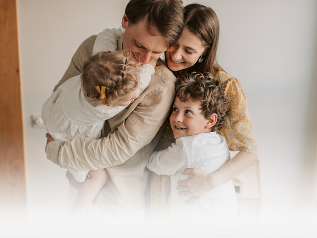A family of four shares a warm, joyful embrace in a brightly lit, indoor setting.