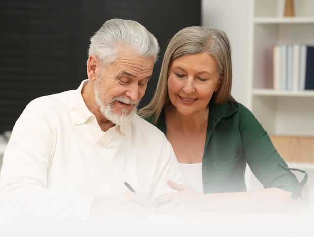 Two people sit at a table looking down at a document while one person signs it with a pen in an office setting.