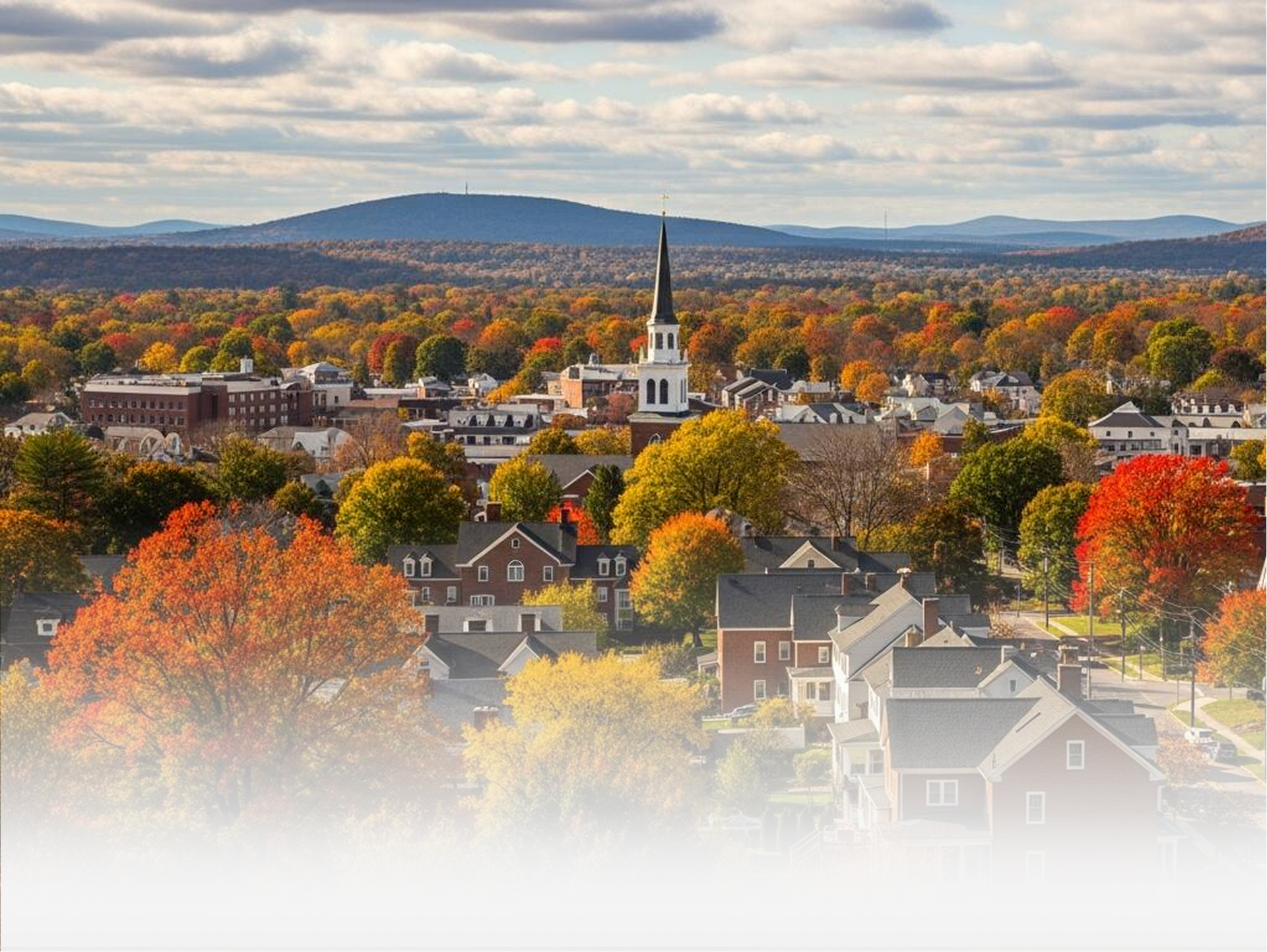 A scenic autumn town view with vibrant fall foliage, historic buildings, a central church steeple, and rolling mountains.
