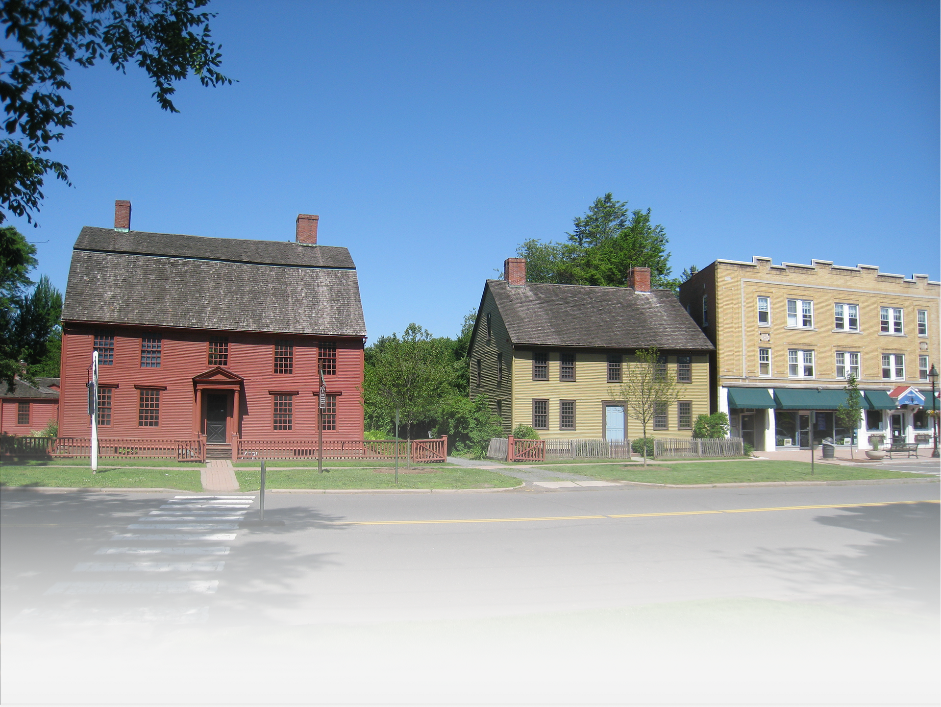 A red colonial-style home, a tan historic house, and a three-story brick commercial building on a street under blue sky.