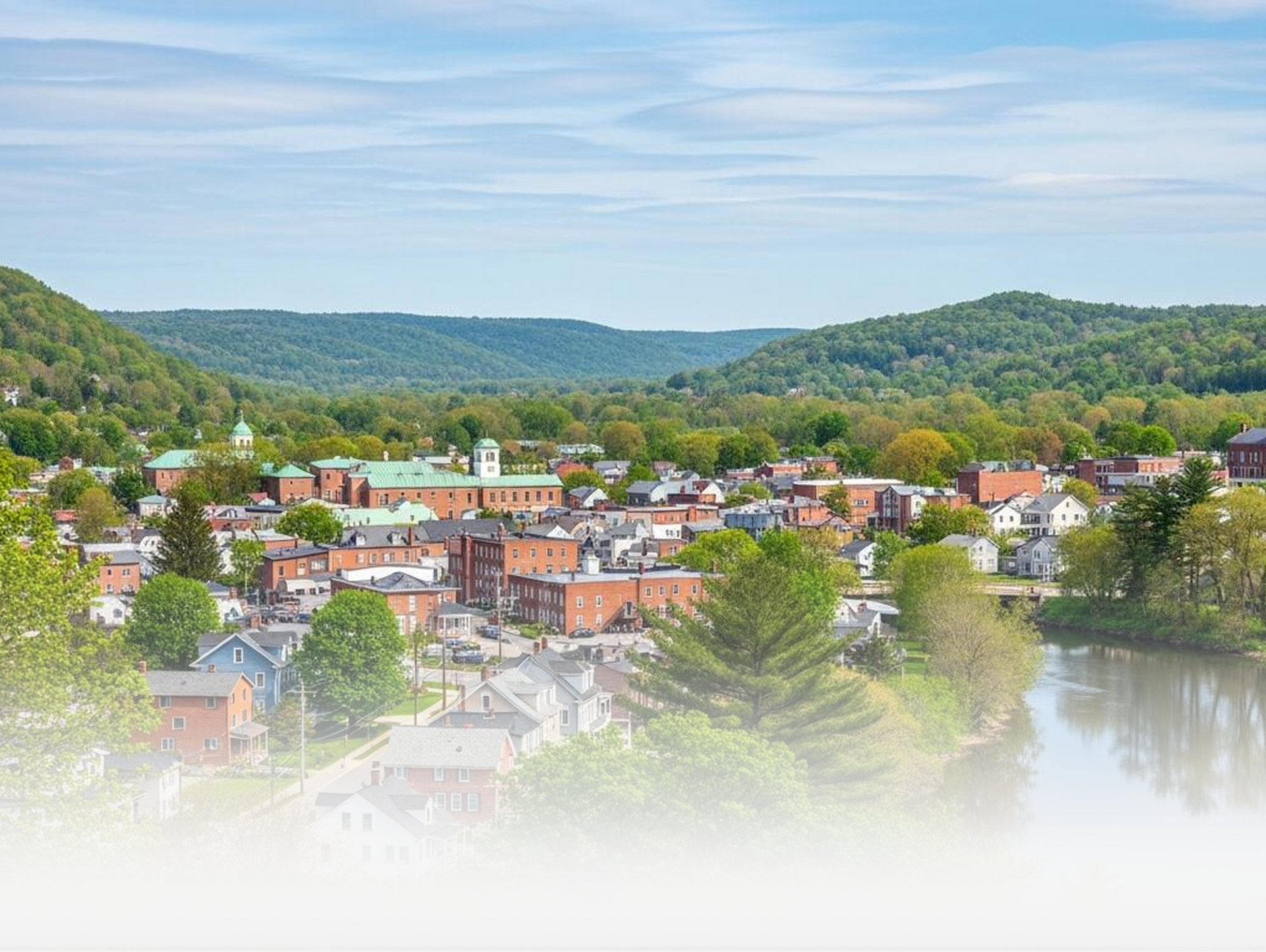 A scenic town nestled in a lush green valley with a river winding alongside, framed by rolling mountains under a blue sky.