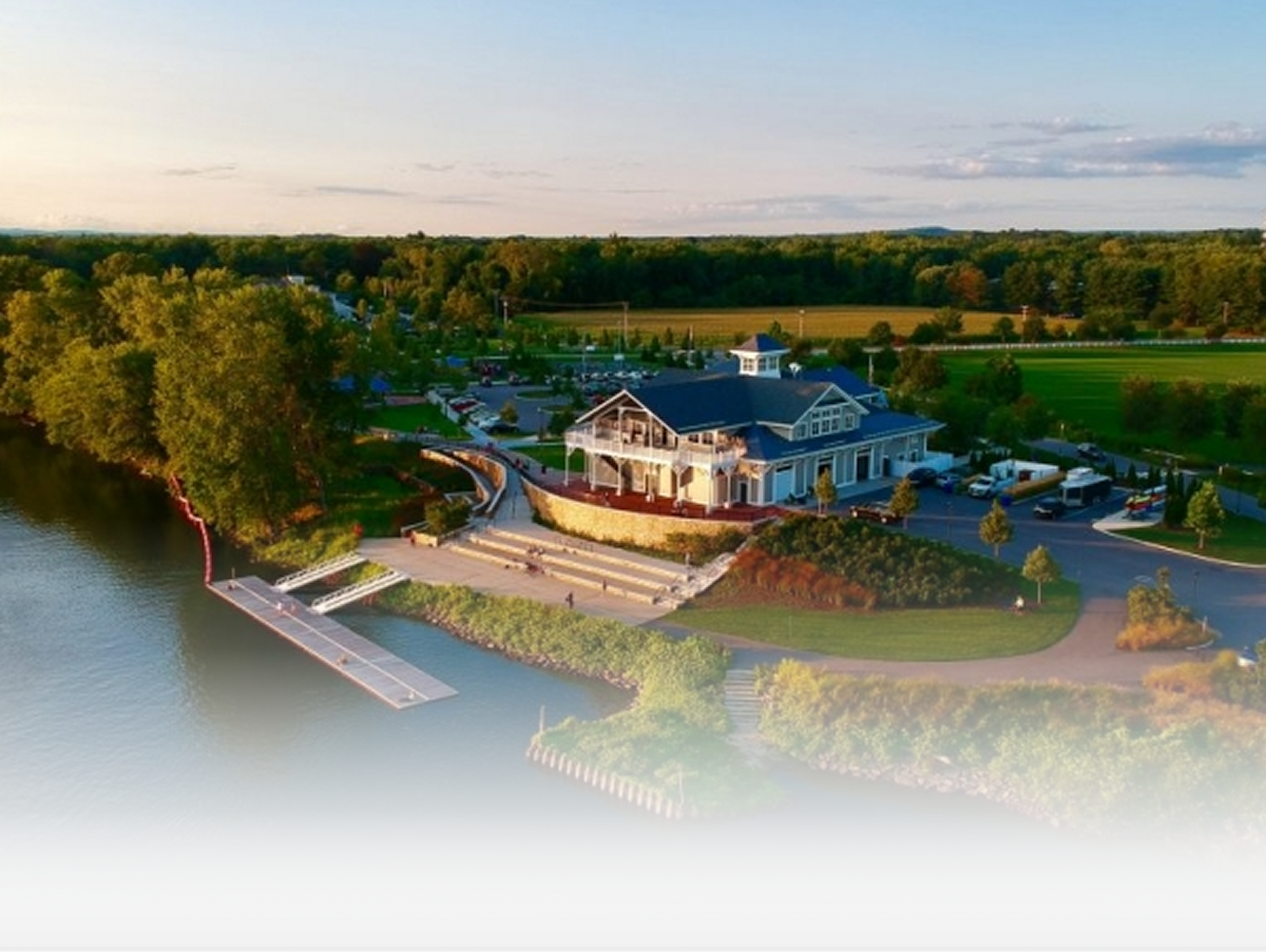 An aerial view of a waterfront building with a tiered patio, dock, and parking area surrounded by trees and green fields.