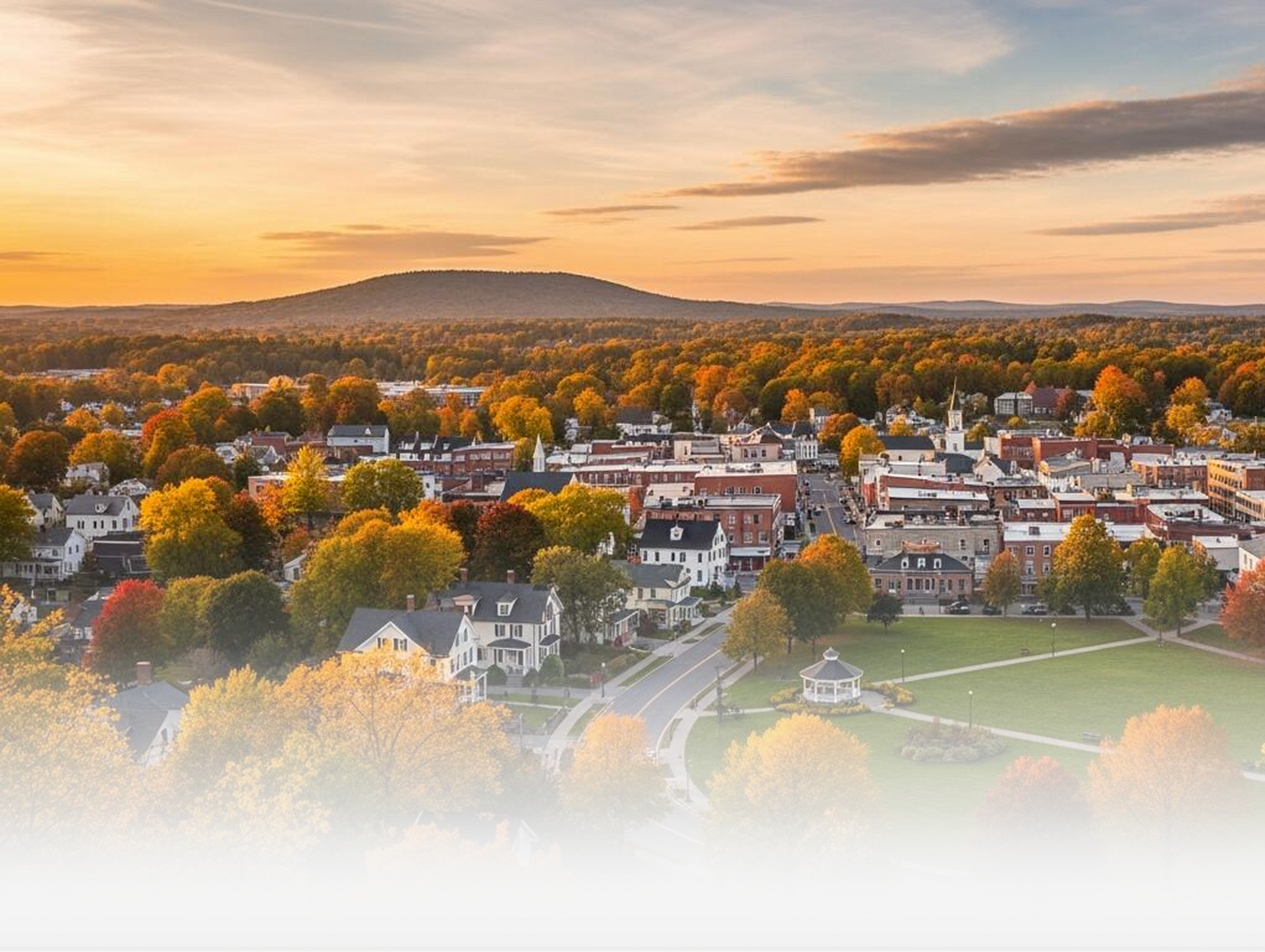 Aerial view of a small town nestled among autumn trees at sunset, with a grassy town green in the foreground.