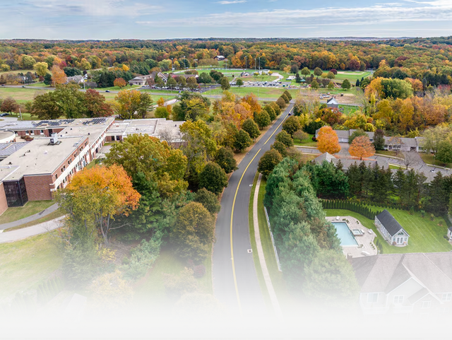 Aerial view of a road winding through a tree-lined landscape with a building on the left and a house with a pool nearby.