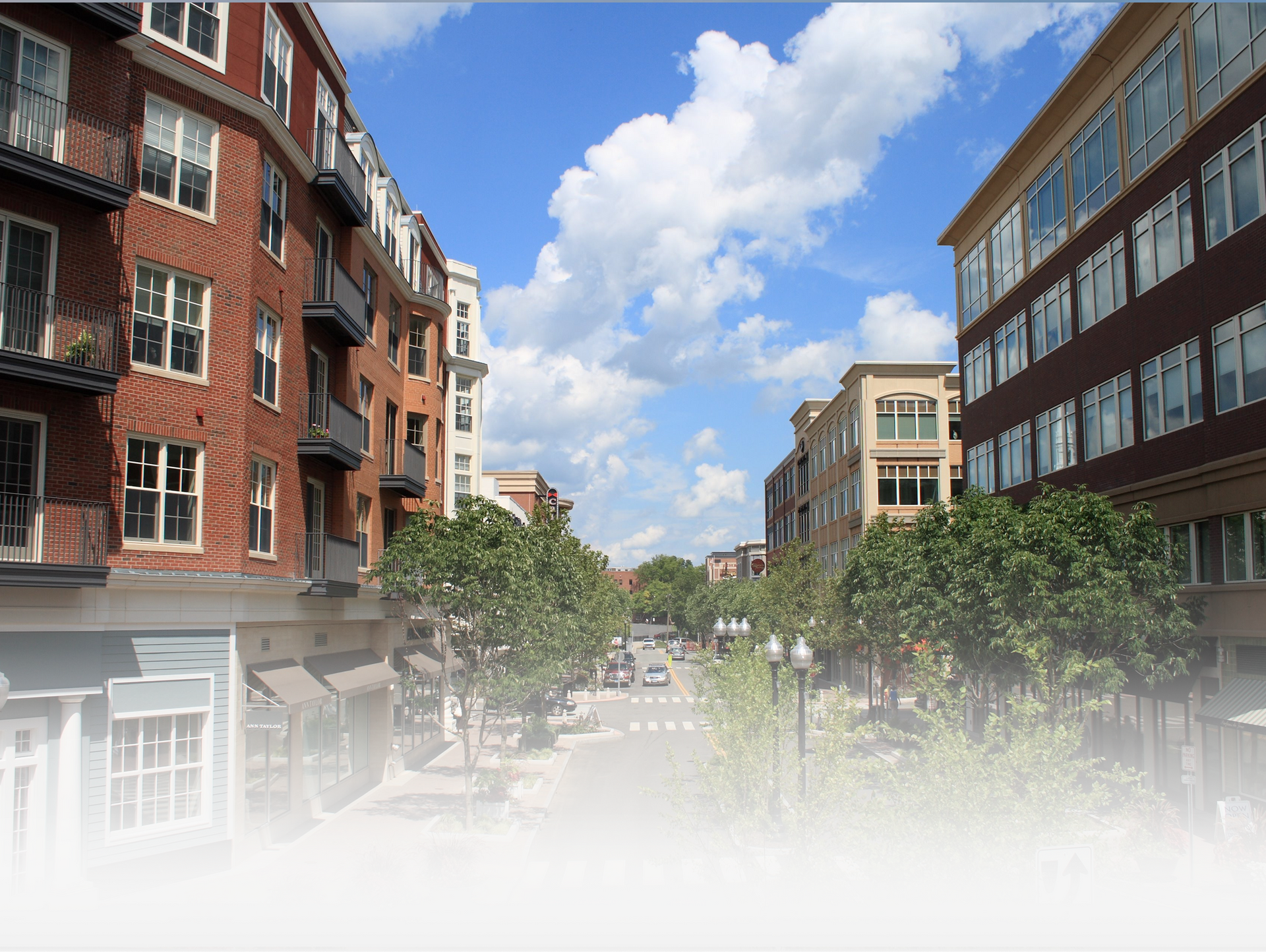 A sunny, tree-lined city street features brick apartment buildings, storefronts, and a clear blue sky with white clouds.