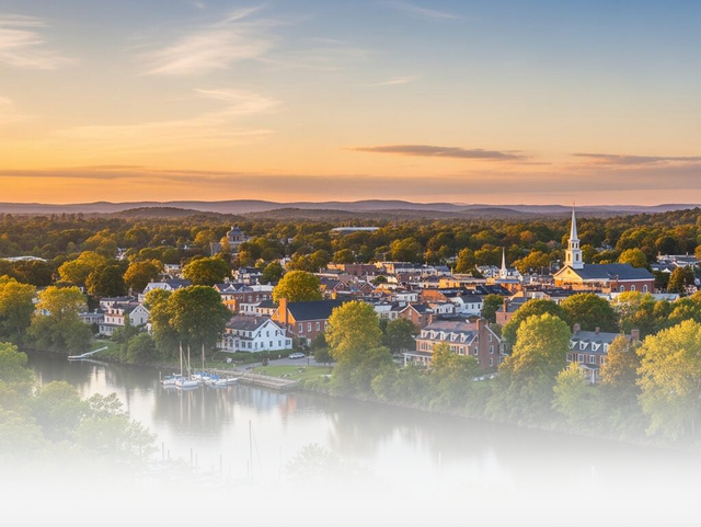 A town with a church steeple sits along a calm river at sunset, with golden light reflecting over the surrounding trees.