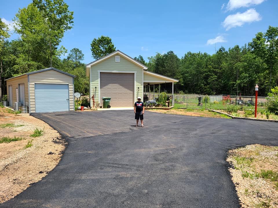 A person stands on a newly paved driveway with two garages in the background, surrounded by trees under a blue sky.