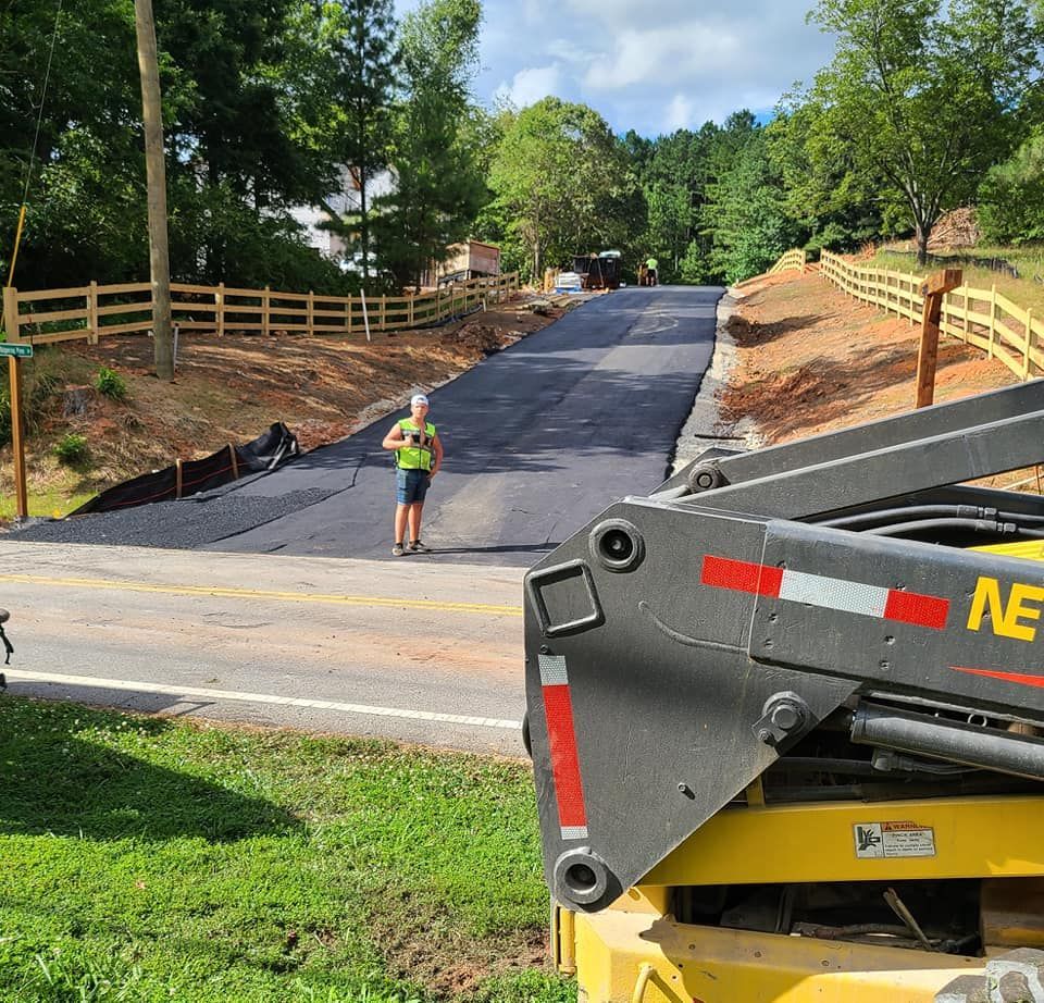 Road construction: paved road uphill. Worker in yellow, fence on sides, machine in foreground. Green grass, trees.