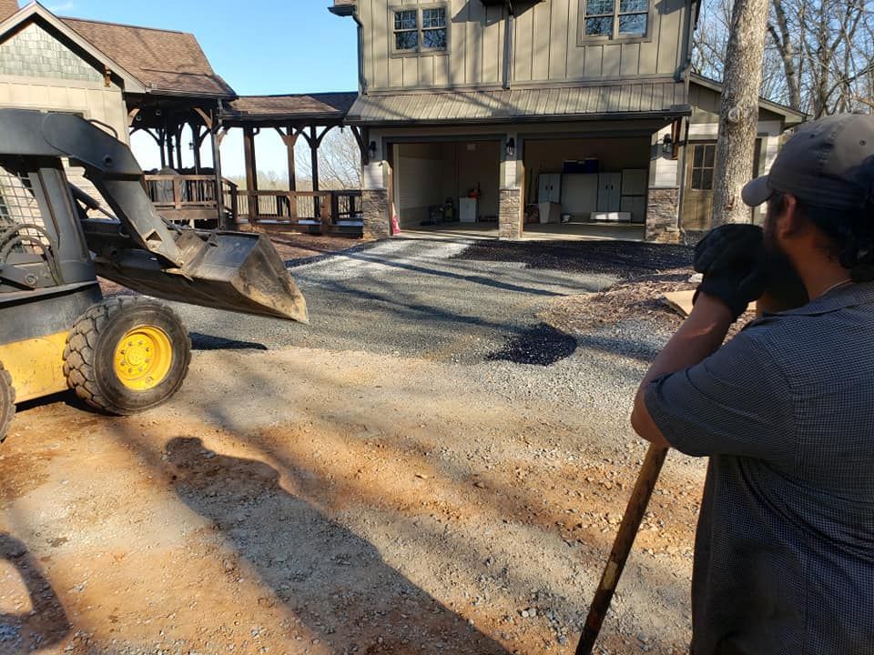 Person holding a shovel watches a skid steer on gravel driveway near a house and covered patio.