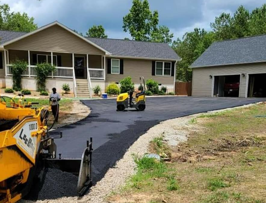 Asphalt driveway being paved, with a house and garage in the background; a worker and paving equipment are visible.
