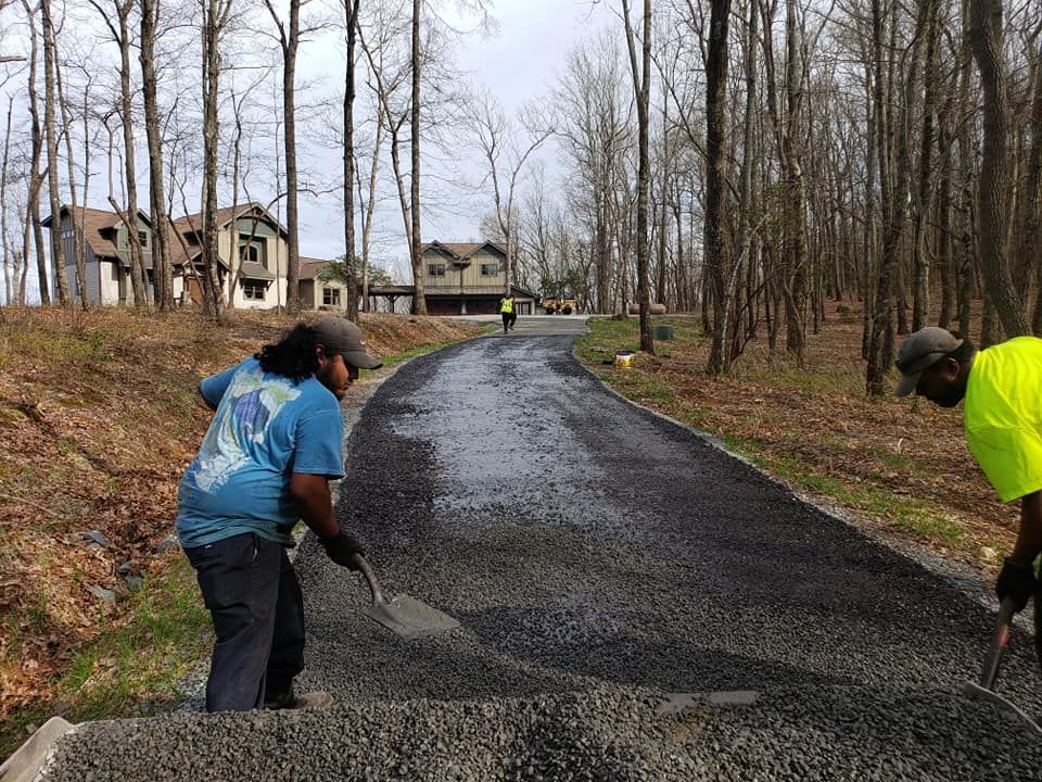 Two workers spreading gravel on a newly paved road in a wooded area near houses.