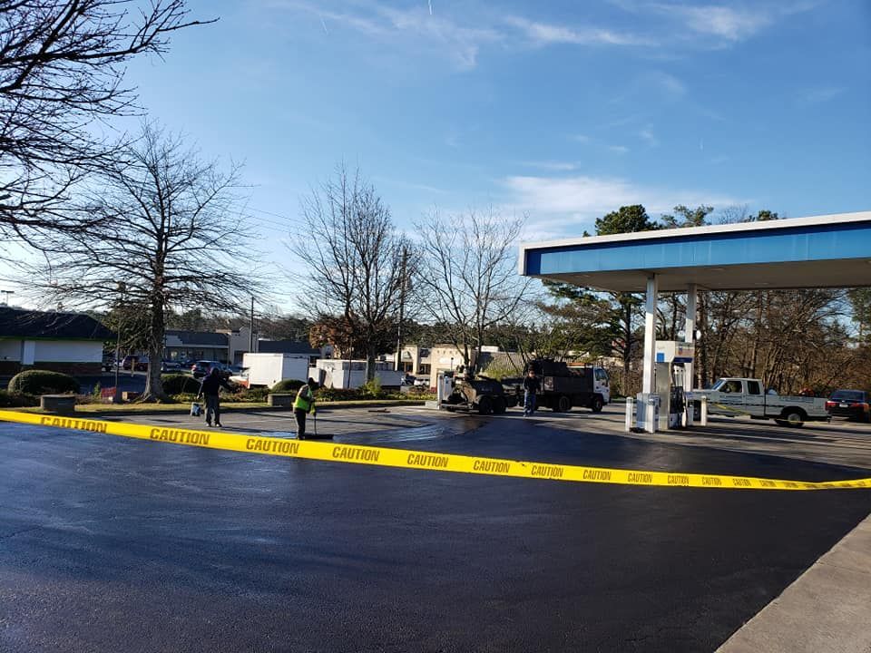 Newly paved asphalt at a gas station with caution tape and workers. Clear sky, trees, and vehicles.