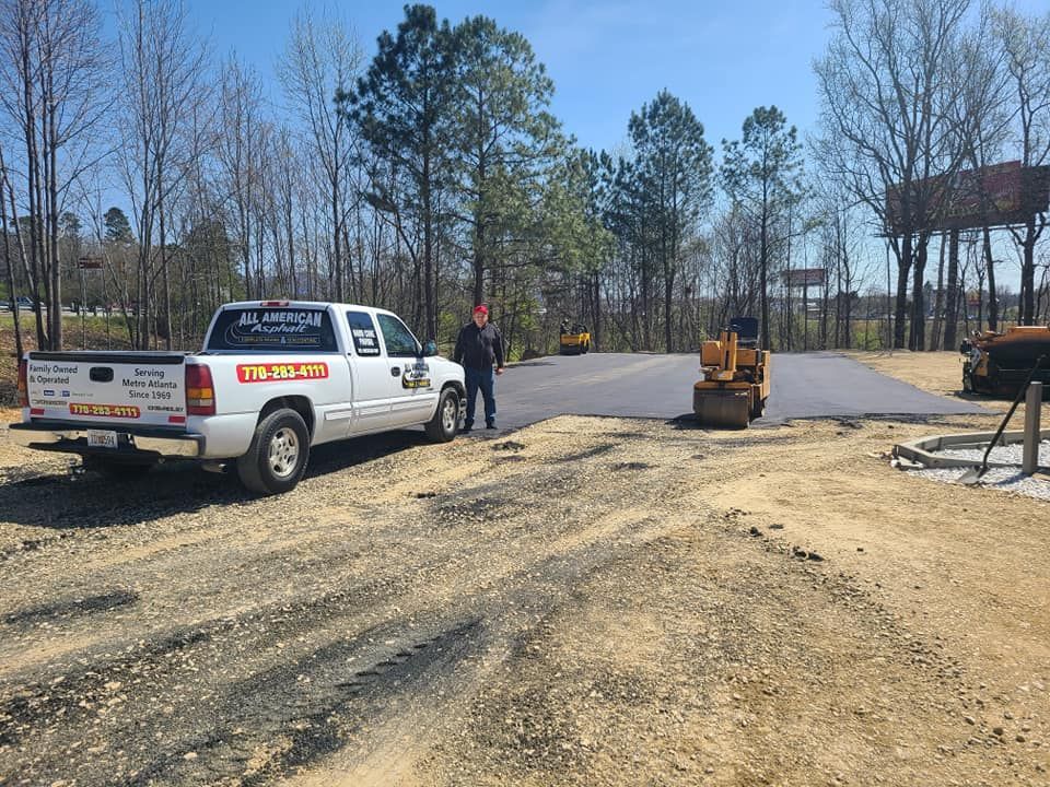 Construction site: worker near white truck, small roller compactor, newly paved asphalt area, dirt foreground, trees.