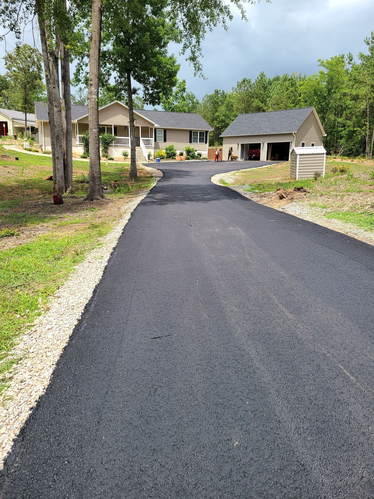 Asphalt driveway leading to a garage under a two-story house with a wooden porch on the left.