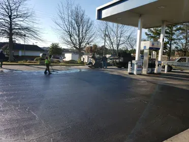 Gas station with wet pavement, several people. One is sweeping. Canopy overhead.