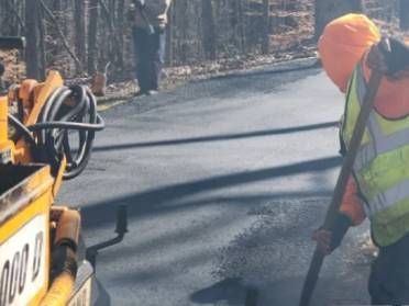 Road workers paving a road. One spreads asphalt with a shovel, another watches.