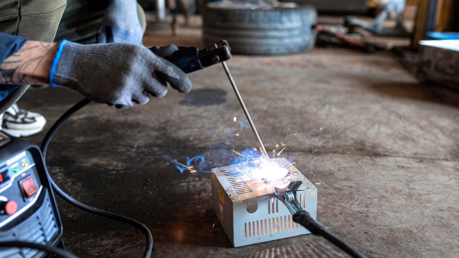 Person welding metal box; sparks fly. Gray gloves, black cable, indoor setting.