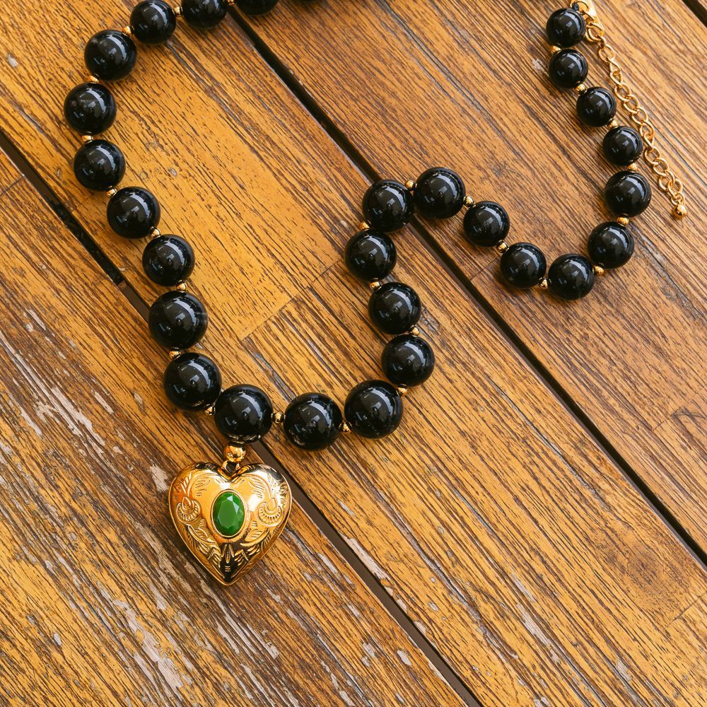 Collar de cuentas negras con un colgante de corazón dorado y una piedra verde sobre una base de madera.