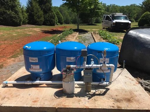 Man operating drilling rig, spraying water and mud. Green trees in background.