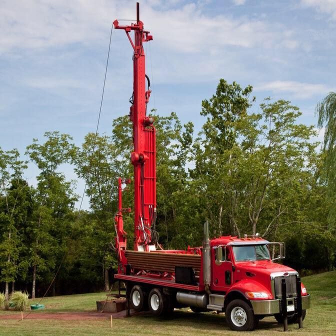 Red water well drilling truck in a grassy field with trees under a blue sky.