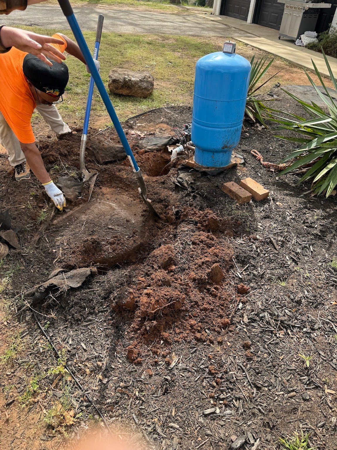 Two people dig around a blue water tank in a yard, removing dirt with shovels.