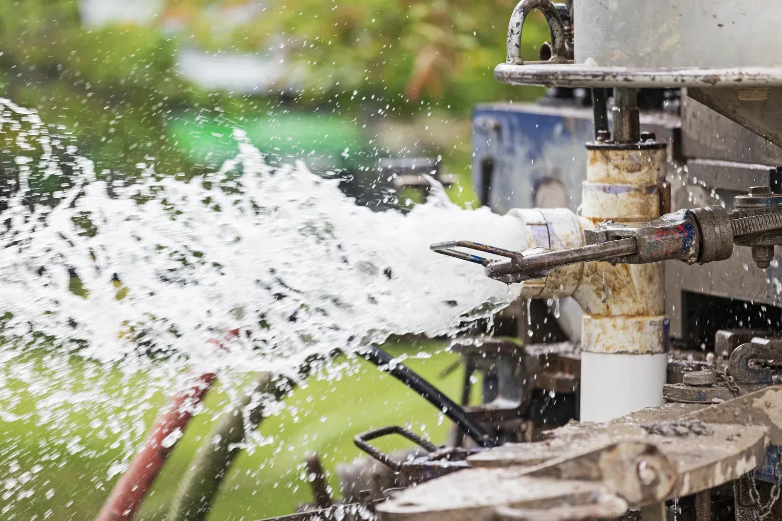 Well pump components: blue tank, exposed pipes, in-ground well and access lid, surrounded by dirt and landscaping.