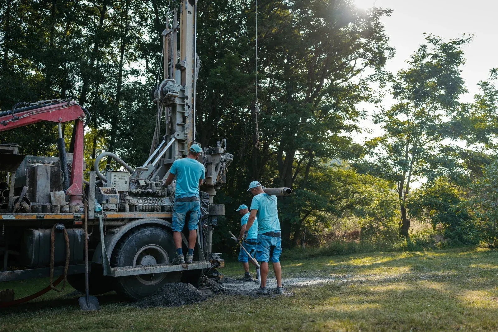 Men in blue shirts operate a drilling rig in a grassy area, sunlight filtering through trees.