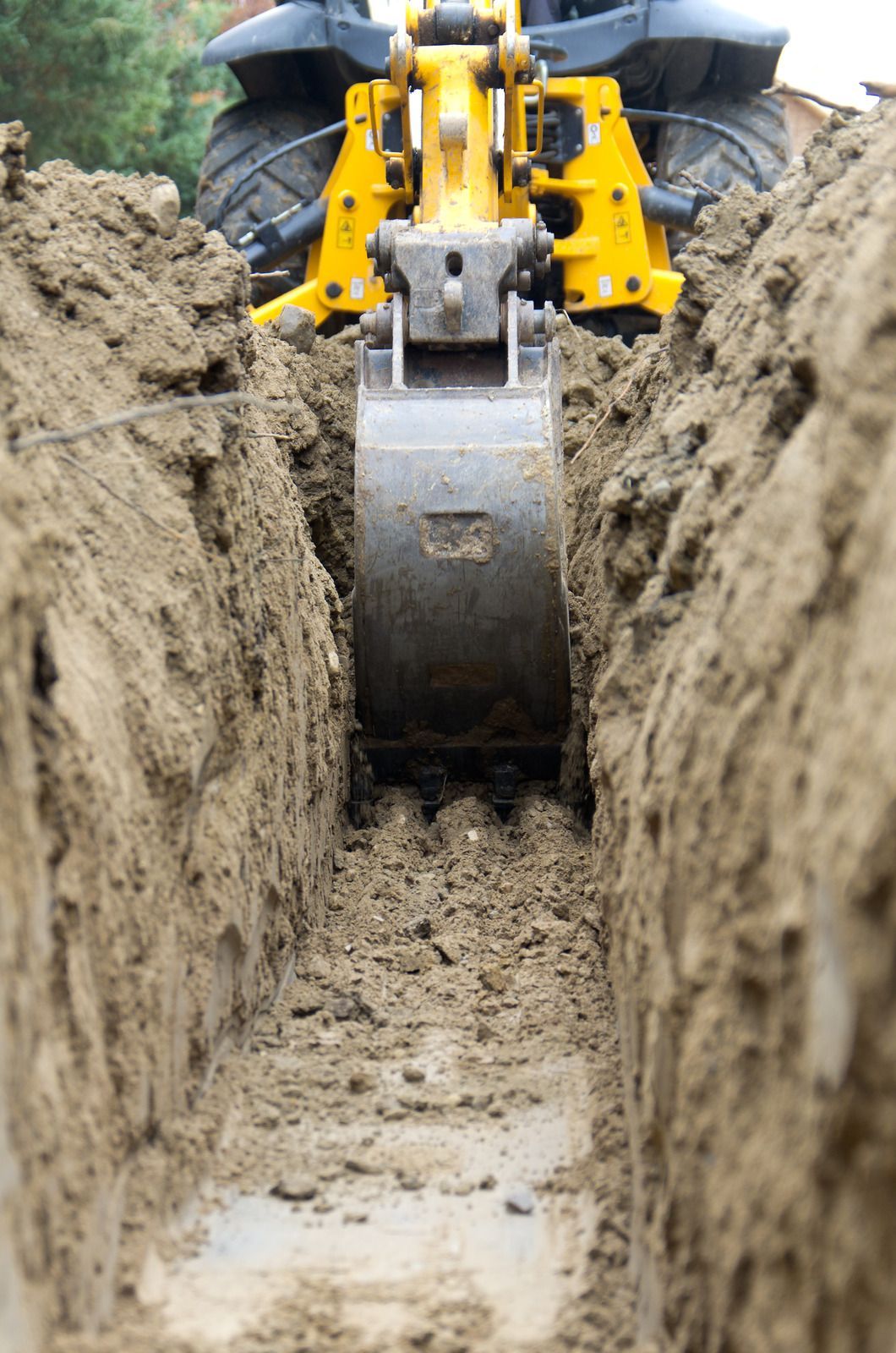 Yellow excavator bucket digging a narrow trench in brown earth.