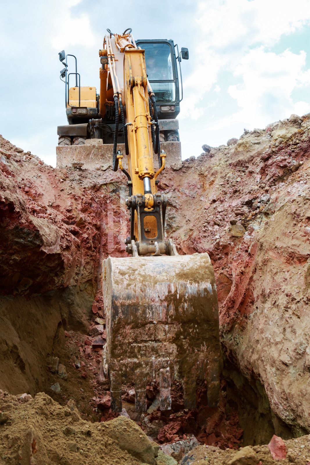 Yellow excavator digging into reddish-brown earth, under a cloudy sky.