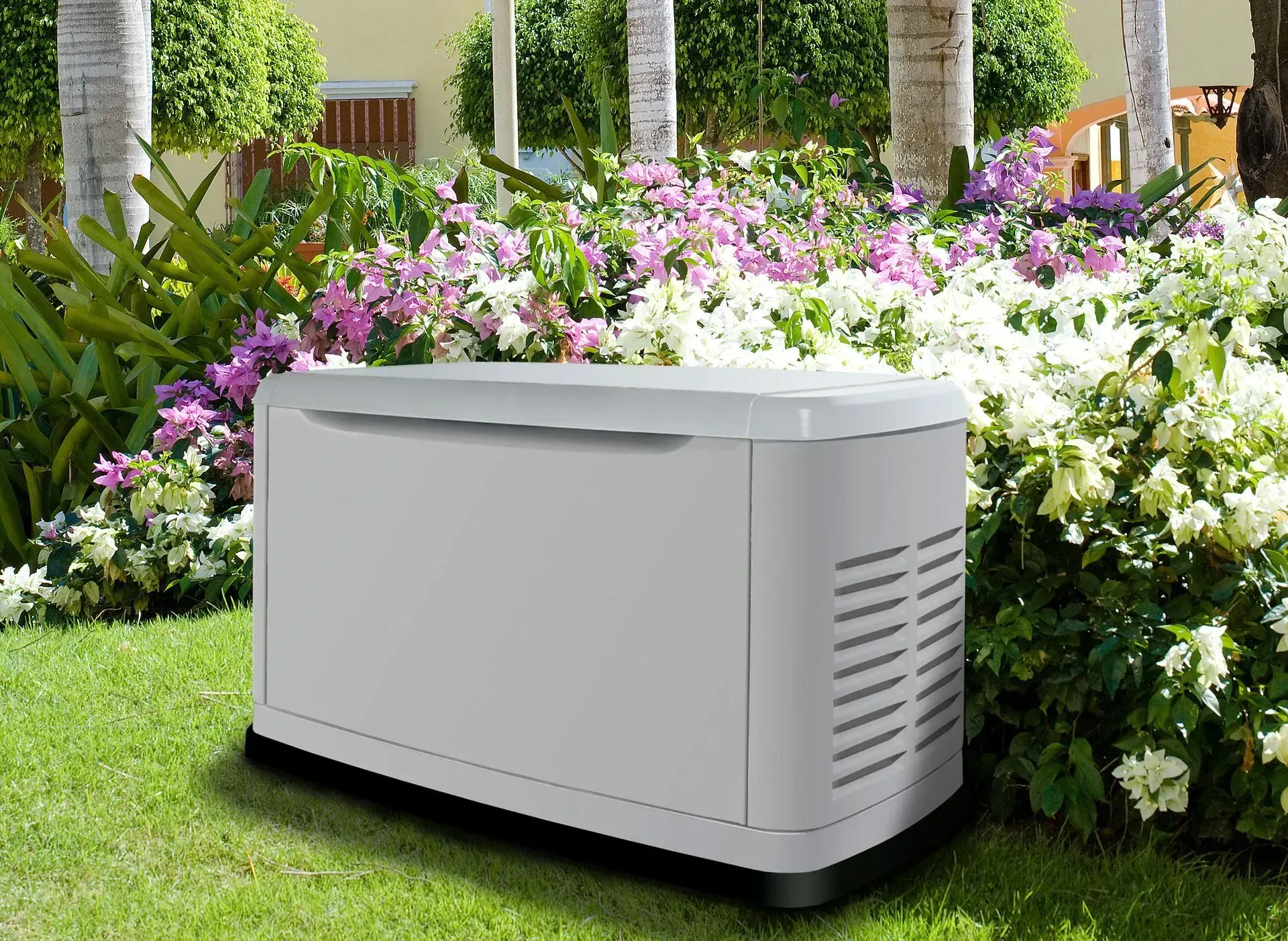 A light gray, rectangular standby generator sits on a lawn in front of a blooming garden with white and purple flowers.