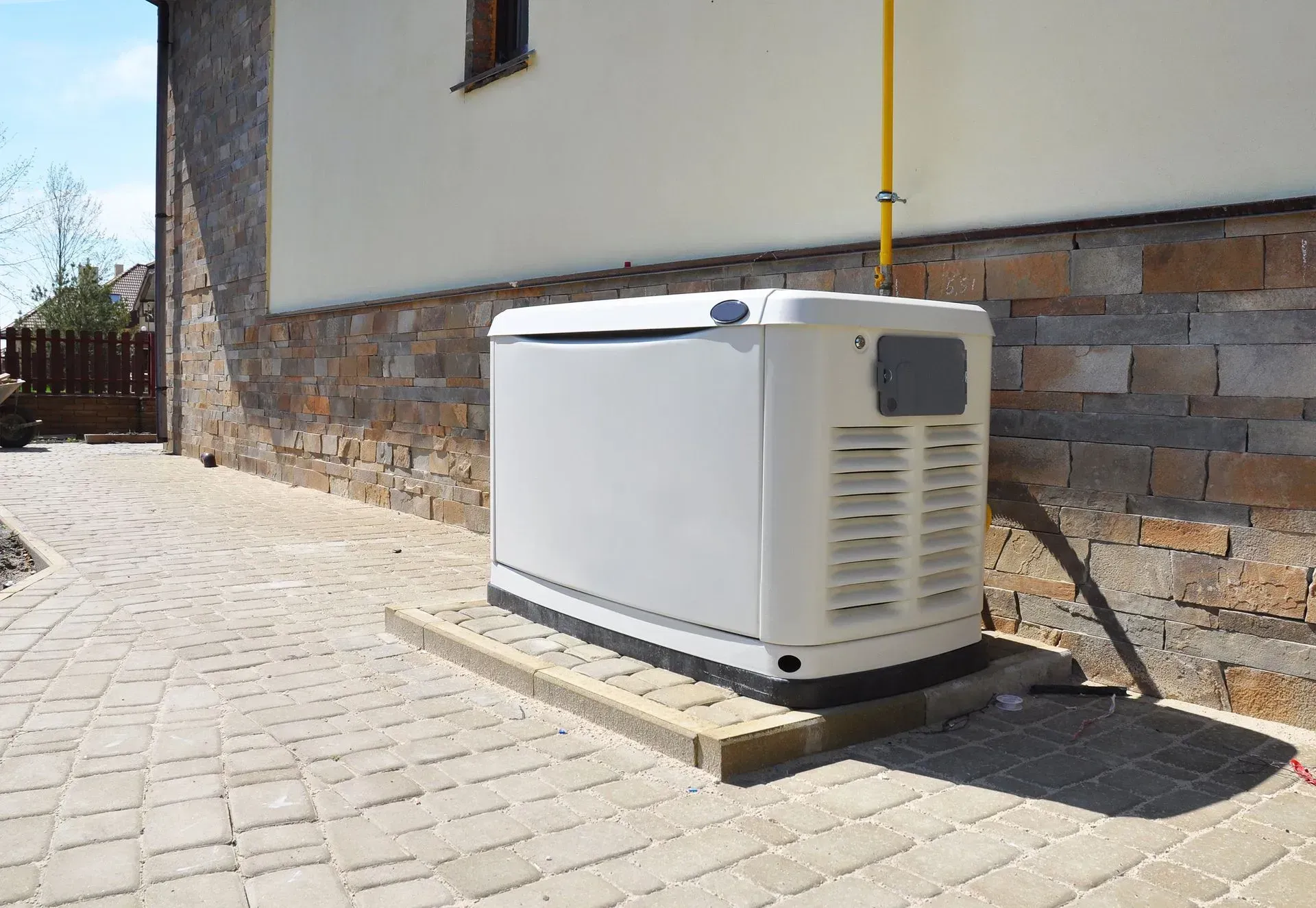 A white residential backup generator sits on a paved stone pad next to a stone and stucco house wall.