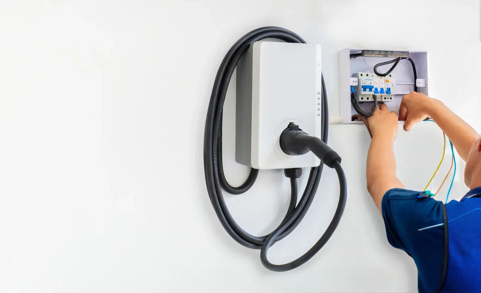 An electrician installing a white electric vehicle charging station on a wall, connecting wires into an open breaker box.