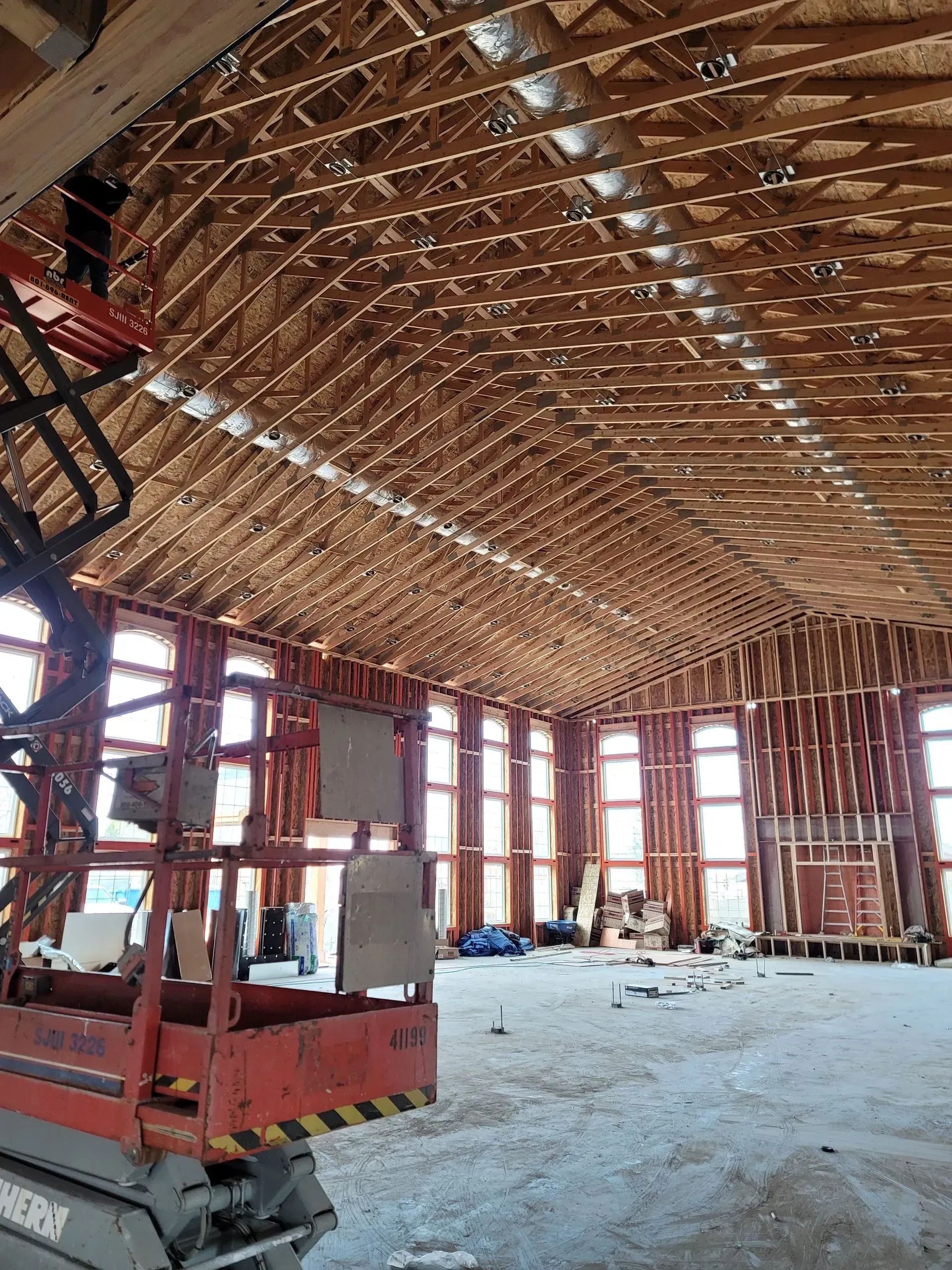 An unfinished building interior featuring a tall vaulted wooden truss ceiling, large window frames, and a red scissor lift.