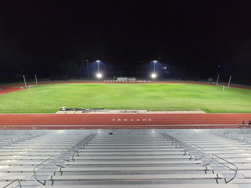 Empty stadium bleachers overlooking a green sports field illuminated by bright floodlights at night.