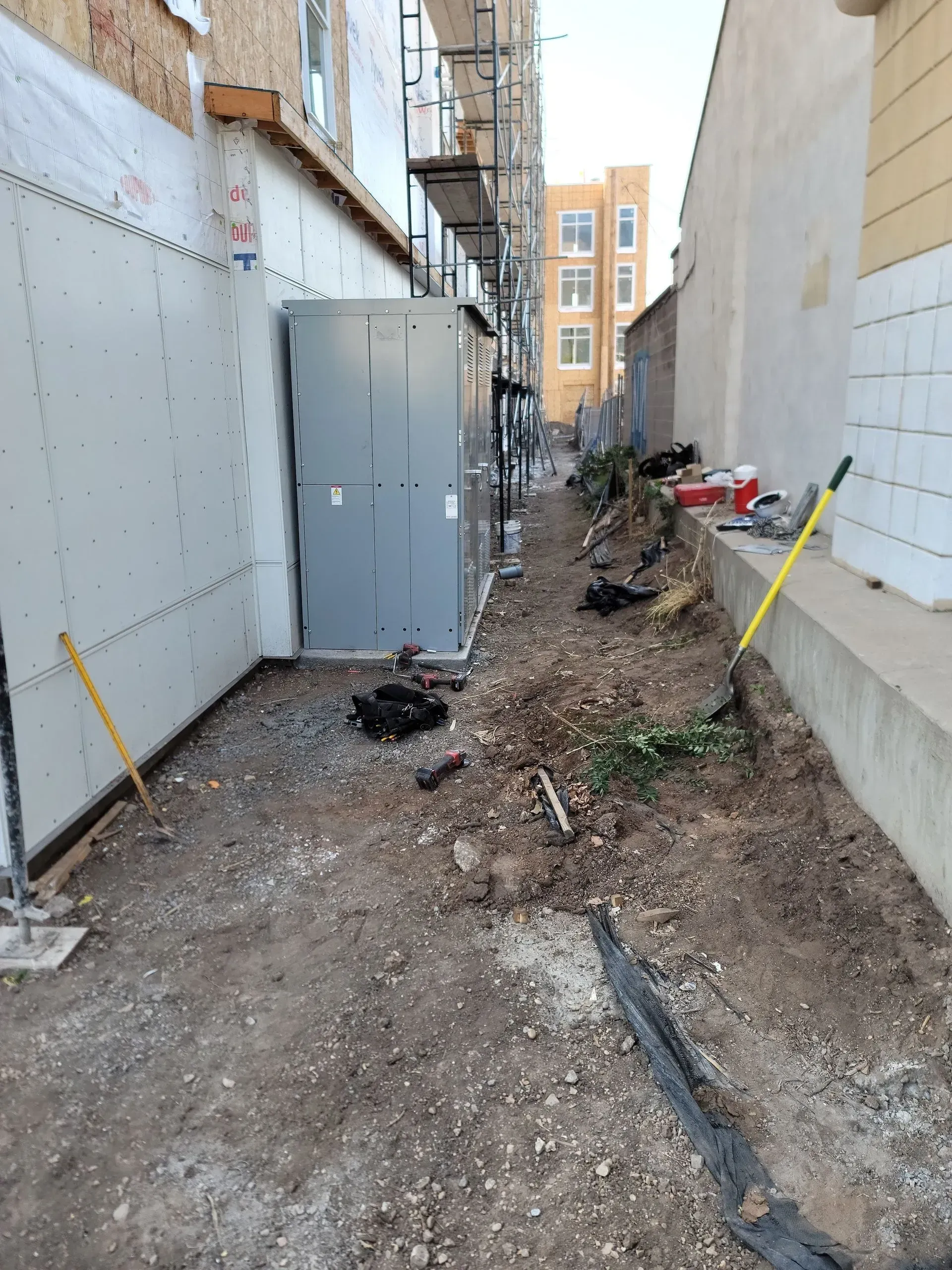 Construction site alleyway with an electrical cabinet, scaffolding, tools on the ground, and a building under development.