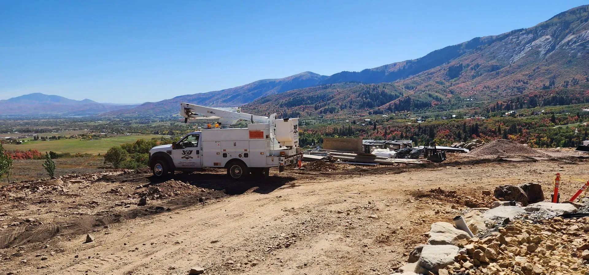 A white utility bucket truck parked on a dirt construction site in front of a mountainous, sunny landscape.